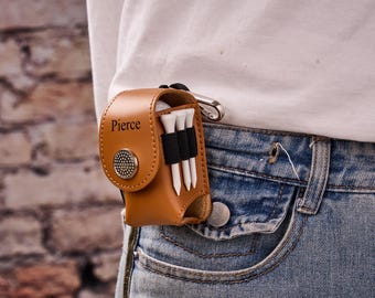 May include: A brown leather golf tee holder with the name "Pierce" printed on the front. The holder is attached to a pair of blue jeans with a silver carabiner clip. Three white golf tees are held in place by a black elastic band.