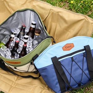 May include: Two insulated cooler bags on a tan blanket on grass. One is green and cream, open, filled with ice and bottles. The other is blue and light blue with black straps and the word "Time" on a brown patch.
