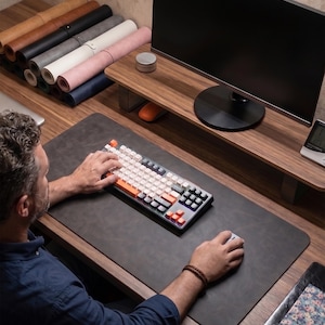 May include: A dark gray rectangular desk mat made of a smooth material, positioned on a wooden desk surface. The mat features rounded corners and provides a workspace for a mechanical keyboard with white and orange keys and a gray computer mouse.