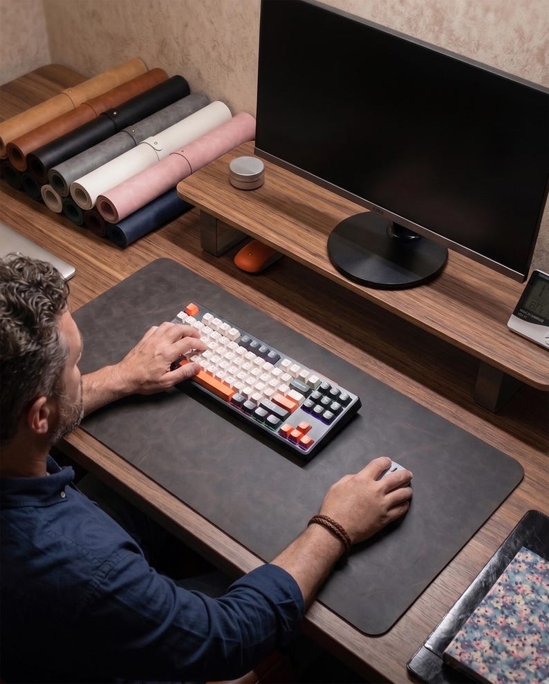 May include: A dark gray rectangular desk mat made of a smooth material, positioned on a wooden desk surface. The mat features rounded corners and provides a workspace for a mechanical keyboard with white and orange keys and a gray computer mouse.