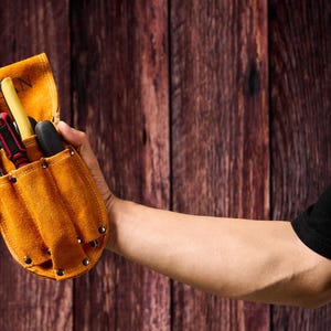 May include: A tan leather tool belt with multiple pockets, filled with various tools including screwdrivers and a wrench. The belt is held in a person's hand against a wooden background.