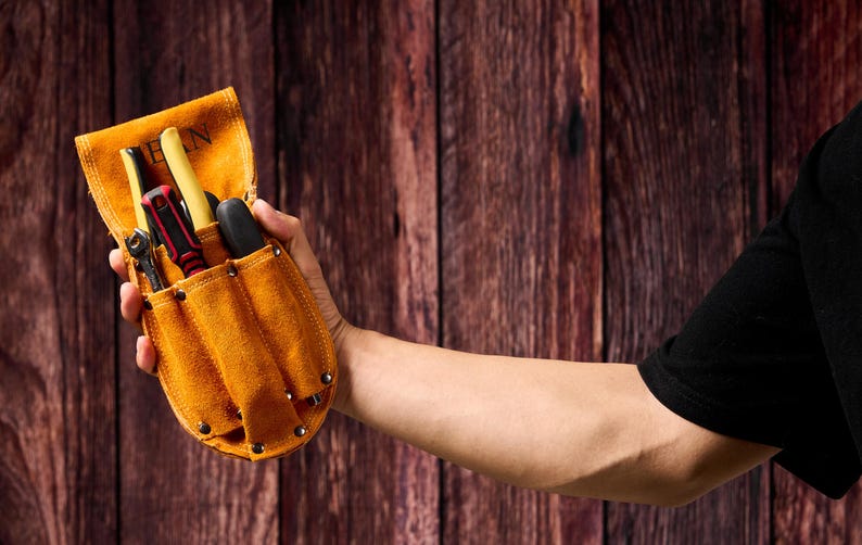May include: A tan leather tool belt with multiple pockets, filled with various tools including screwdrivers and a wrench. The belt is held in a person's hand against a wooden background.