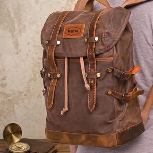 Könnte beinhalten: Ein brauner Rucksack aus gewachstem Segeltuch mit hellbraunen Lederriemen, messingfarbenen Metallschnallen und einem Lederaufnäher mit der Prägung RMR. Der Rucksack verfügt über einen Kordelzug, seitliche Kompressionsriemen und einen verstärkten Boden.