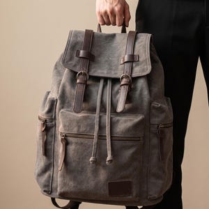 May include: A charcoal gray backpack featuring a top flap with two brown leather straps and metal buckles. The bag includes a front pocket with a horizontal zipper, two side pockets with zippers, and a drawstring closure under the main flap.