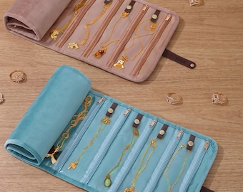 May include: Two velvet jewelry rolls, one dusty rose and one teal, each featuring multiple internal zippered compartments and snap-button tabs. Gold necklaces are displayed inside the rolls, while several gold rings are scattered nearby.