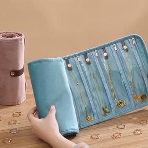 May include: A dusty rose velvet jewelry roll with a brown strap and gold snap closure, alongside an open teal blue jewelry organizer featuring four clear zippered compartments containing gold necklaces and several gold rings scattered nearby.