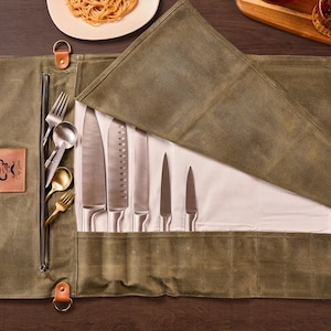 Peut inclure: Un étui à couteaux en toile vert olive ouvert, contenant divers couteaux et couverts en acier inoxydable. Le rouleau est partiellement déroulé, révélant les couteaux et les ustensiles. Un patch en cuir avec un logo est visible.