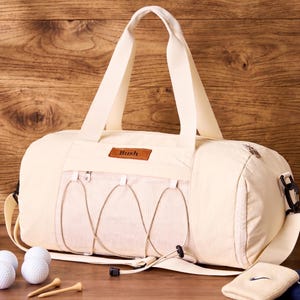 Può includere: Borsa da viaggio color crema con una tasca frontale e la scritta "Bush" su una toppa in pelle marrone. La borsa ha una tracolla e un manico superiore. Palline e tee da golf in primo piano.