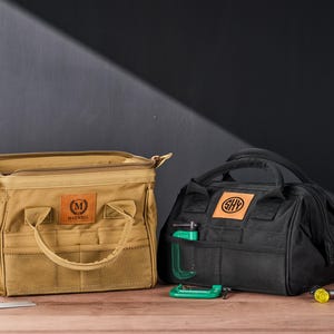 May include: Two tool bags, one tan and one black, sit on a wooden surface. The tan bag has the name "Maxwell" on a leather patch. The black bag has the initials "SHY" on a leather patch. A green clamp is in the black bag's front pocket.