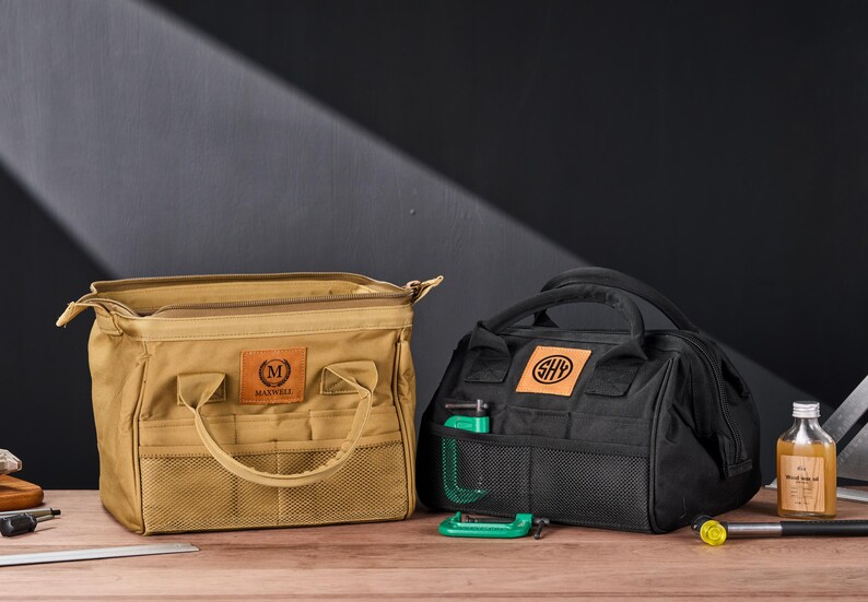 May include: Two tool bags, one tan and one black, sit on a wooden surface. The tan bag has the name "Maxwell" on a leather patch. The black bag has the initials "SHY" on a leather patch. A green clamp is in the black bag's front pocket.