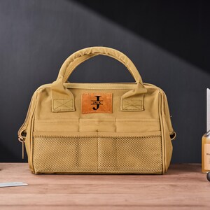 May include: Tan canvas tool bag with a brown leather patch that reads "JACK" with the initial "J". The bag has a handle, mesh pockets, and a zipper closure. A bottle of wood wax oil and a metal square are in the background.
