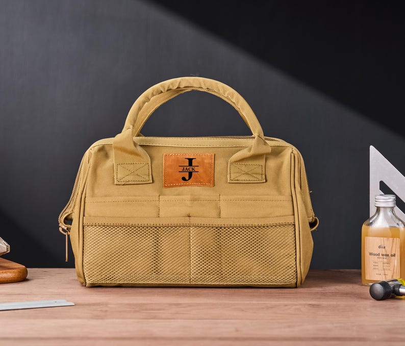 May include: Tan canvas tool bag with a brown leather patch that reads "JACK" with the initial "J". The bag has a handle, mesh pockets, and a zipper closure. A bottle of wood wax oil and a metal square are in the background.