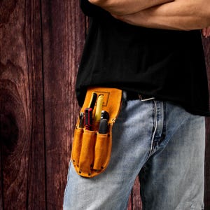 May include: A tan leather tool belt with several pockets, worn at the waist. The belt contains various tools, such as screwdrivers and pliers. The person is wearing a black t-shirt and blue jeans.