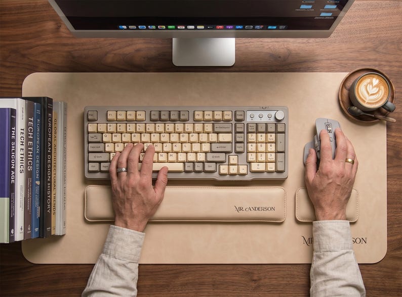 May include: A beige leather desk mat featuring the text MR. CANDERSON, paired with a matching beige leather wrist rest. The set includes a grey mechanical keyboard with cream-colored keys and a grey computer mouse, all arranged on a wooden surface.