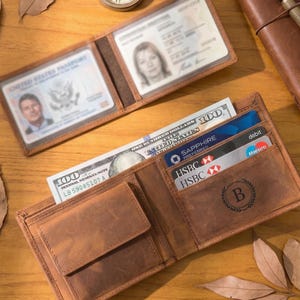 May include: Open brown leather wallet with multiple card slots, a bill compartment holding a $100 bill, and a small coin pocket. The wallet also holds a passport and an ID card. The wallet is embossed with the letter "B".