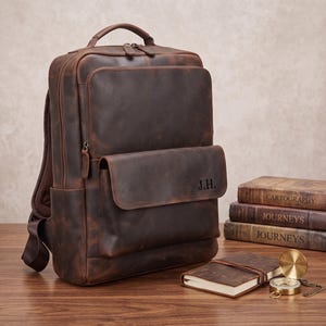 Könnte beinhalten: Ein dunkelbrauner Lederrucksack mit Tragegriff, einem großen Hauptfach und einer Fronttasche mit Klappe, auf der die Initialen J.H. eingeprägt sind. Der Rucksack verfügt über sichtbare Nähte, Metallreißverschlüsse und verstellbare Schultergurte.