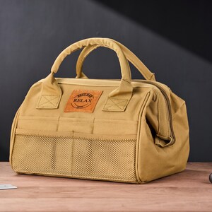 May include: A tan canvas tool bag with a top handle and a front mesh pocket. The bag has a brown leather patch with the text "BEST DAD RELAX 1977". The bag is sitting on a wooden surface.