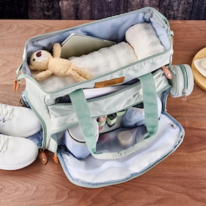 以下が含まれることがあります： ぬいぐるみ、タブレット、タオルなどが入った、明るい緑と青のトラベルバッグ。白いスニーカーがバッグの横にあります。その他のアイテムには、小さな丸い容器、石鹸とボトルが入った木製のトレイなどがあります。