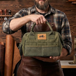May include: An olive green tool bag with a handle and multiple pockets, held by a person. The bag features a rectangular patch with a mountain logo and the word "SOURCE". The bag is made of a durable material and is designed for carrying tools.