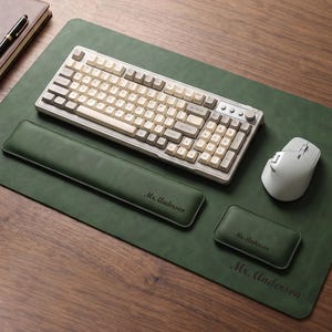May include: A dark green desk mat with a matching wrist rest and mouse pad, all featuring the name Mr. Anderson. A mechanical keyboard with cream and grey keys and a white wireless mouse are placed on the mat, which has a smooth, matte finish.