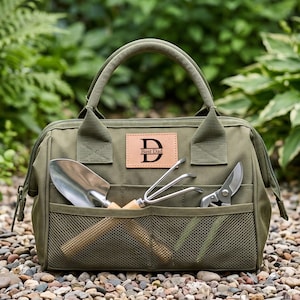 May include: An olive green canvas garden tool bag with two top handles and a front mesh pocket. A tan leather patch on the front features a large letter D and the text Best Dad. The bag contains a metal hand trowel, a metal hand rake, and pruners.