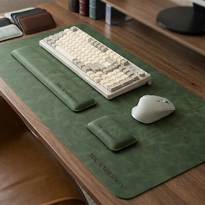 May include: A dark green desk mat with the text MR. ANDERSON, featuring a matching long wrist rest and a small square coaster, both also embossed with MR. ANDERSON. A beige mechanical keyboard and a white wireless mouse sit on the mat.
