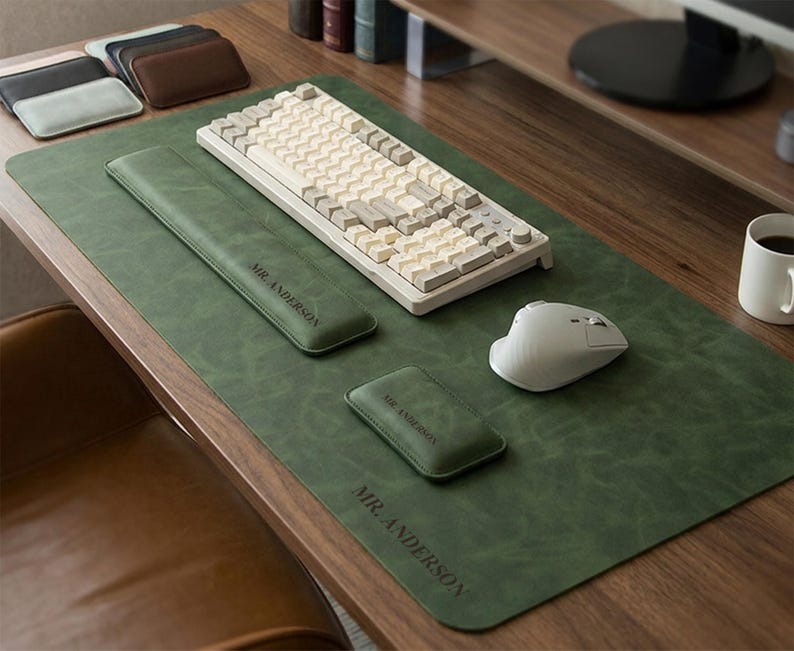 May include: A dark green desk mat with the text MR. ANDERSON, featuring a matching long wrist rest and a small square coaster, both also embossed with MR. ANDERSON. A beige mechanical keyboard and a white wireless mouse sit on the mat.