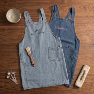 May include: Two denim aprons with cross-back straps and front pockets. The lighter blue apron features the embroidered text "Chef in training" and a wooden-handled brush in the pocket. The darker blue apron features the embroidered text "Hammer".