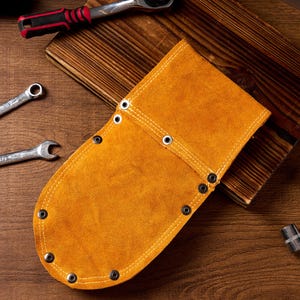 May include: A tan leather tool holder with metal eyelets and stitching. The holder is on a wooden surface with spanners and other tools.