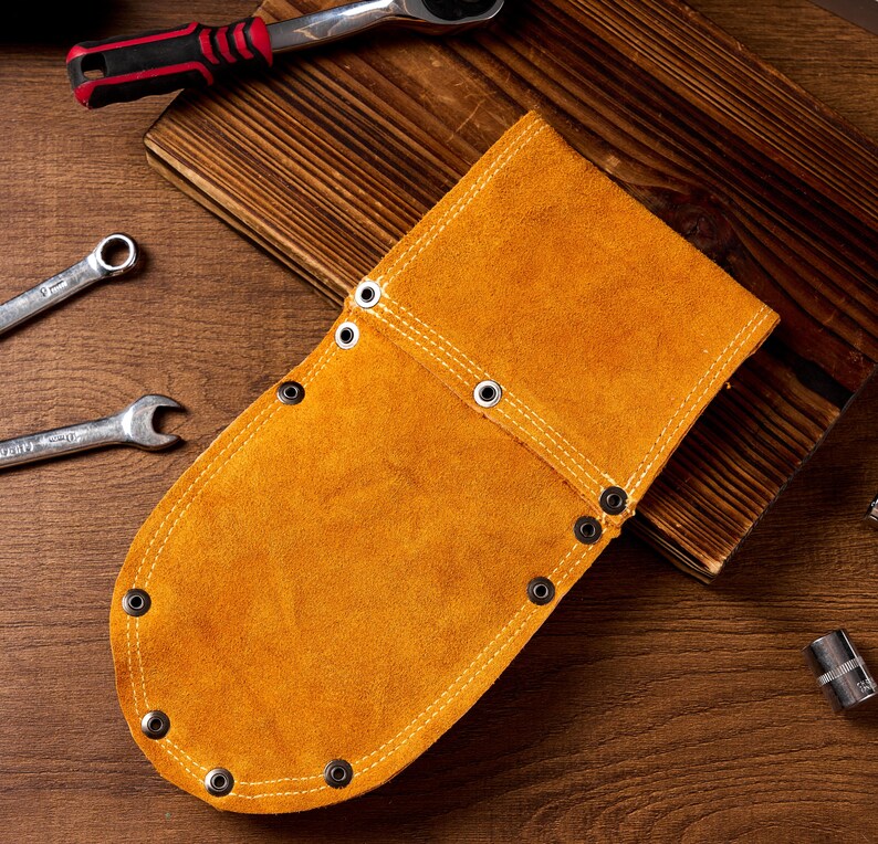 May include: A tan leather tool holder with metal eyelets and stitching. The holder is on a wooden surface with spanners and other tools.
