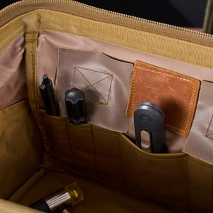 May include: Tan tactical bag with multiple pockets and compartments. The interior features various tools, including a pen, a small hammer, and a knife. The bag has a brown leather patch and is designed for organization and storage.