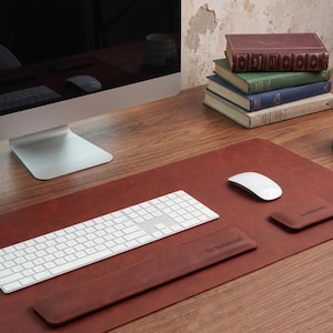 May include: A brown leather desk mat featuring a matching keyboard wrist rest and a small rectangular mouse pad, both embossed with the text The Mastermind. The set is arranged on a wooden surface with a white keyboard and a white computer mouse.