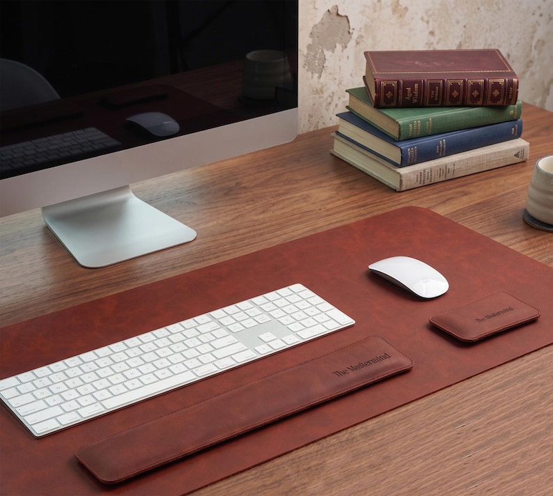 May include: A brown leather desk mat featuring a matching keyboard wrist rest and a small rectangular mouse pad, both embossed with the text The Mastermind. The set is arranged on a wooden surface with a white keyboard and a white computer mouse.