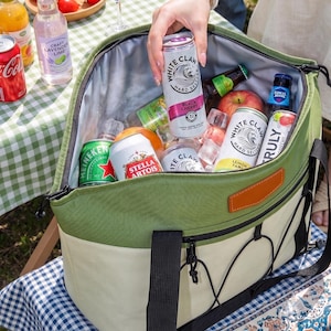 Könnte beinhalten: Eine grüne und beige isolierte Kühltasche gefüllt mit verschiedenen Getränkedosen und Eis. Eine Hand hält eine Dose White Claw Hard Seltzer in der Geschmacksrichtung Black Cherry. Weitere Dosen sind Heineken, Stella Artois und Truly.