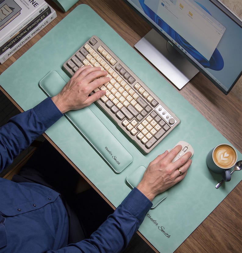 May include: A light green desk mat featuring a matching keyboard wrist rest and mouse wrist rest. The mat and wrist rests are made of a smooth material with the text "Alexander Smith" embossed on the surface. A mechanical keyboard with beige and grey keys sits on the mat.