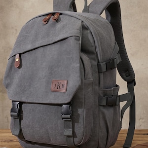 Puede incluir: Una mochila de lona gris con detalles en cuero marrón y hebillas negras. La mochila tiene un cierre de solapa, correas ajustables y un pequeño parche de cuero marrón con las iniciales "JKM".