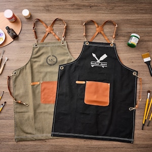 May include: Two aprons, one olive green and one black, featuring brown leather cross-back straps with metal hardware. Each apron has a single brown leather pocket and metal grommets. The olive apron displays the letters MC, while the black apron features a paintbrush graphic and the text Rachael Mears.