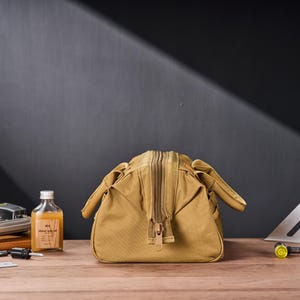 May include: A tan canvas bag with a large zipper closure. The bag has a rectangular shape with rounded sides and small handles. A small bottle of liquid with a label is visible to the left.