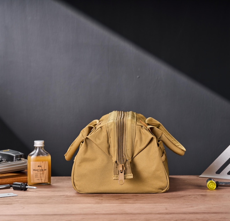 May include: A tan canvas bag with a large zipper closure. The bag has a rectangular shape with rounded sides and small handles. A small bottle of liquid with a label is visible to the left.