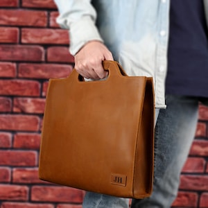 May include: A brown leather tote bag with a rectangular shape and a handle cut-out at the top. The bag has the initials "JH" embossed on the lower right corner. The bag is held by a person.