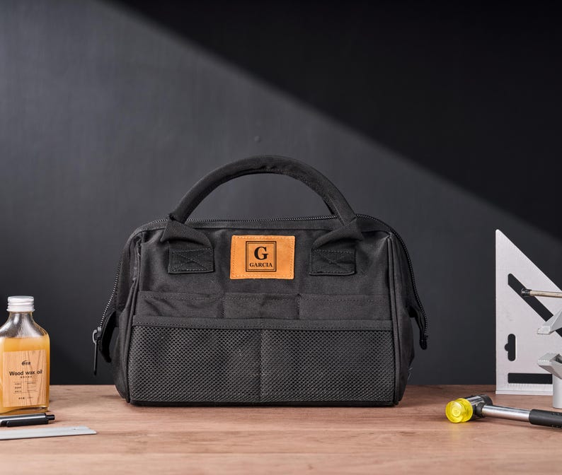 May include: Black tool bag with a handle, front mesh pockets, and a leather patch with the letter "G" and the name "GARCIA". The bag is on a wooden surface with a bottle of liquid, a pen, and a small square.
