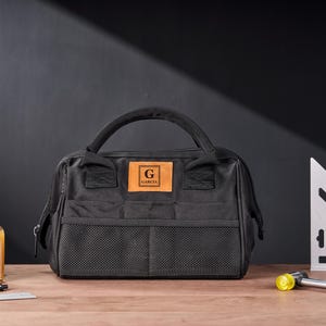 May include: Black tool bag with a handle, front mesh pockets, and a leather patch with the letter "G" and the name "GARCIA". The bag is on a wooden surface with a bottle of liquid, a pen, and a small square.