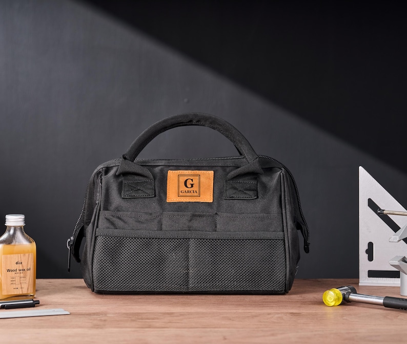 May include: Black tool bag with a handle, front mesh pockets, and a leather patch with the letter "G" and the name "GARCIA". The bag is on a wooden surface with a bottle of liquid, a pen, and a small square.