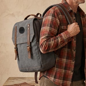 May include: A grey canvas backpack featuring brown leather straps with metal buckles, a top handle, and a circular black leather patch embossed with the name TOM. The backpack has a fold-over flap design and adjustable shoulder straps.