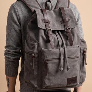 Può includere: Uno zaino in tela grigia con cinturini in pelle marrone scuro con fibbie in metallo, chiusura a coulisse e due tasche laterali. La parte anteriore include una tasca con cerniera e una toppa in pelle marrone con il testo Patricia.