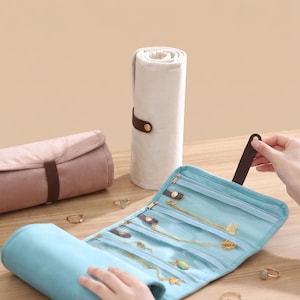 May include: A light blue jewelry roll organizer featuring multiple clear zippered compartments containing gold necklaces and pendants, with a dark brown strap and snap closure. A dusty pink roll and a cream-colored roll are also visible.
