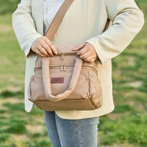 Op de afbeelding: Een bruin schoudertasje met een handvat aan de bovenkant en een crossbody-riem. De tas heeft een gewatteerd ontwerp aan de zijkanten en een kleine rechthoekige patch met de tekst "Lucy". De tas wordt vastgehouden door een persoon die een crèmekleurige blazer draagt.