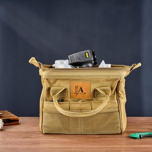 May include: A tan canvas tool bag with multiple pockets and a handle. The bag is open, revealing tools inside. A leather patch with the letter "A" is on the front. The bag is on a wooden surface.