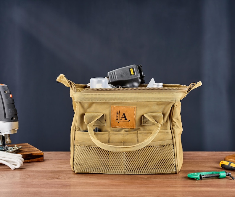 May include: A tan canvas tool bag with multiple pockets and a handle. The bag is open, revealing tools inside. A leather patch with the letter "A" is on the front. The bag is on a wooden surface.