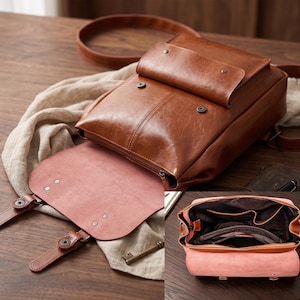 May include: A brown leather backpack with a flap closure and a smaller front pocket. The interior is visible, revealing multiple compartments. The bag has a shoulder strap and a pink interior lining. The bag is shown open and closed.